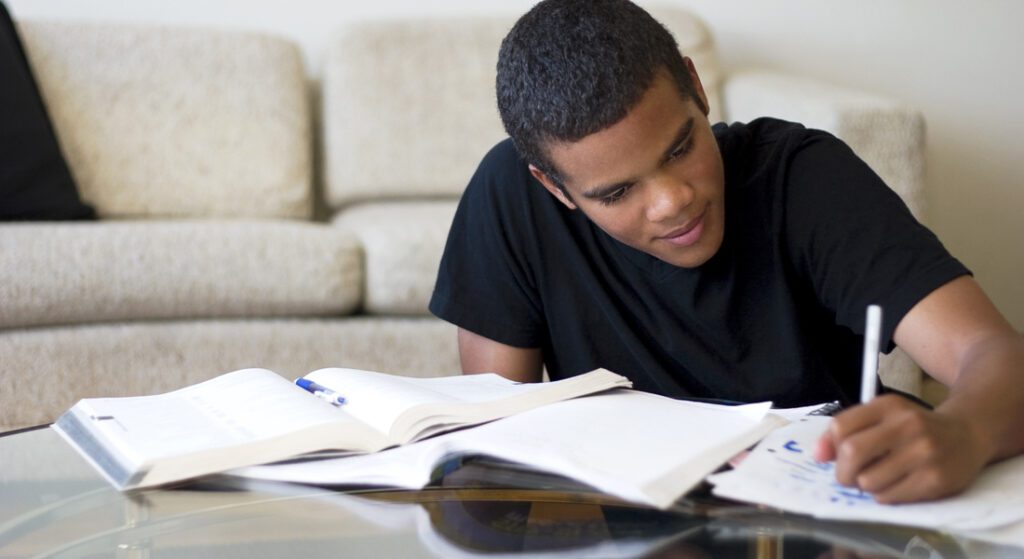 Teenage boy writing on table with books and paper spread around him